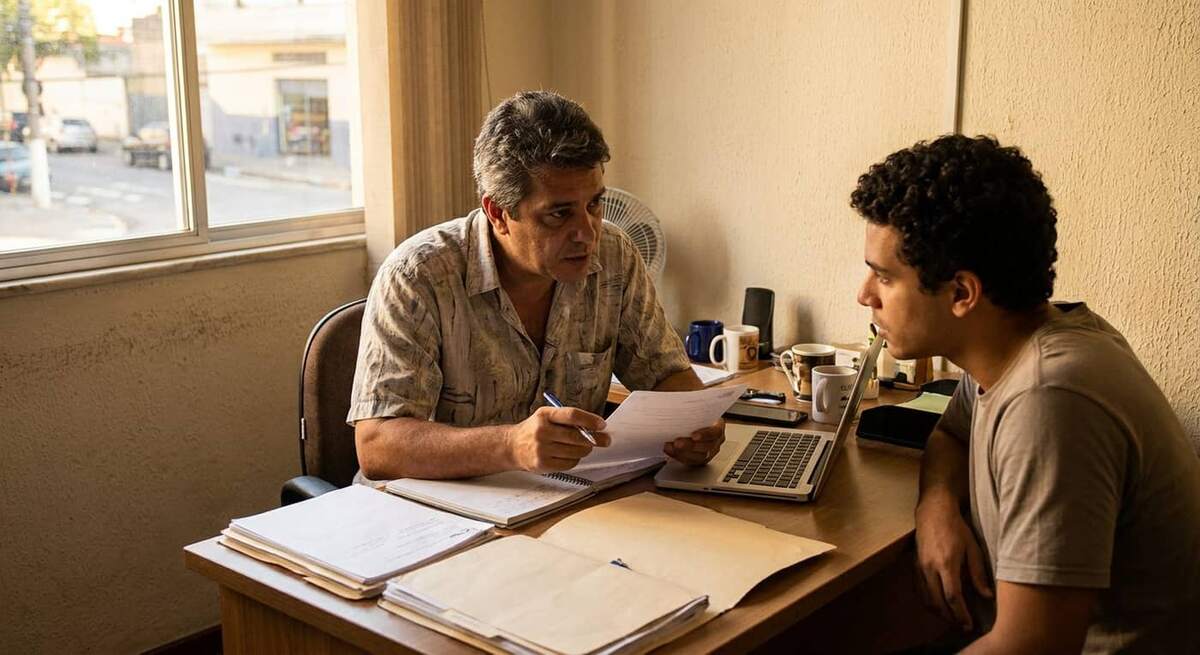 Trabalhador uniformizado recebendo contrato intermitente assinado do dono de pequena empresa em balcão de atendimento com luz natural