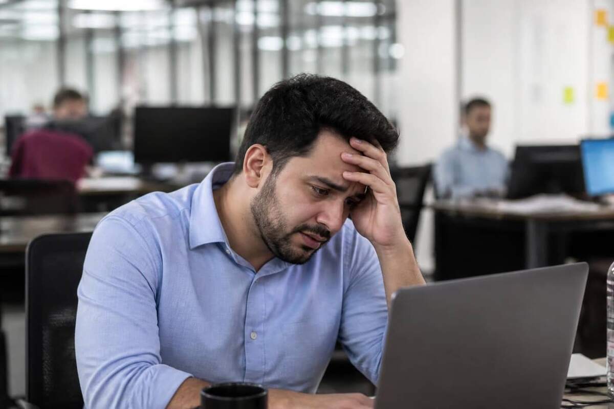 Profissional brasileiro recém-promovido a gestor sentado em escritório moderno olhando para o laptop com expressão de esgotamento e mão na cabeça